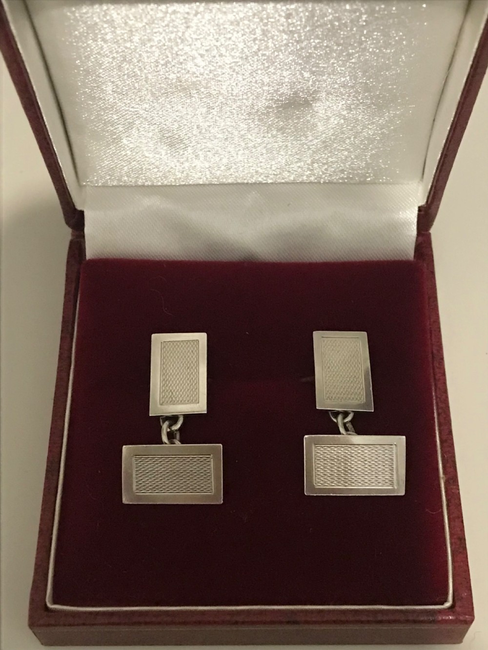 A pair of rectangular silver engine-turned cufflinks connected by chains, displayed inside an open fitted jewellers box. View III