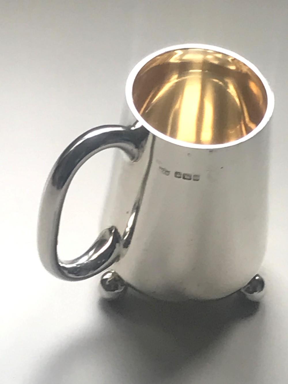 An Edwardian sterling silver christening mug featuring a tapered body, loop handle, gold gilt interior, ball feet, and visible hallmarking.