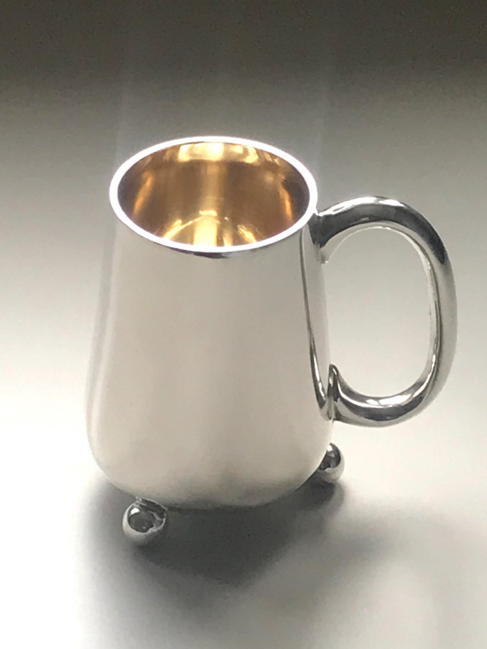 An Edwardian sterling silver christening mug featuring a tapered body, loop handle, gold gilt interior, ball feet, and visible hallmarking. View II