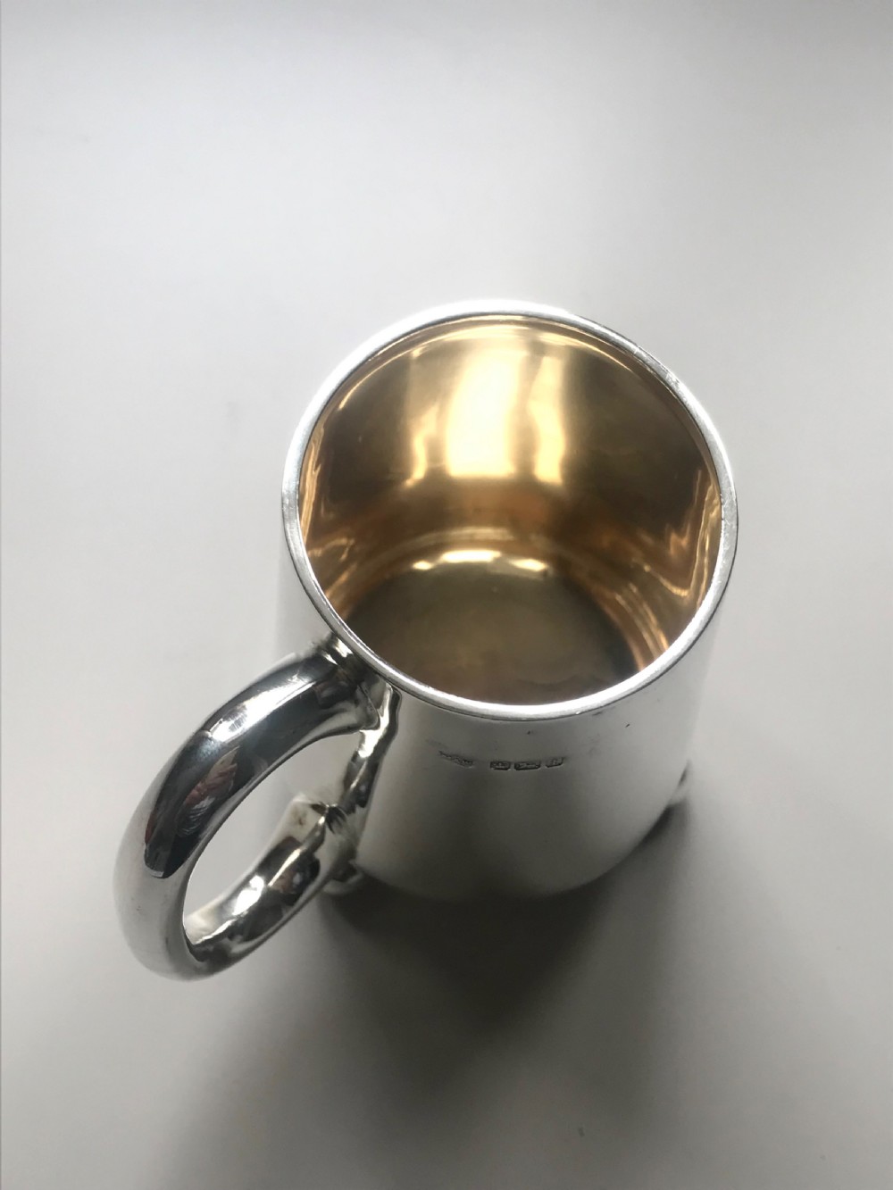 An Edwardian sterling silver christening mug featuring a tapered body, loop handle, gold gilt interior, ball feet, and visible hallmarking. View V