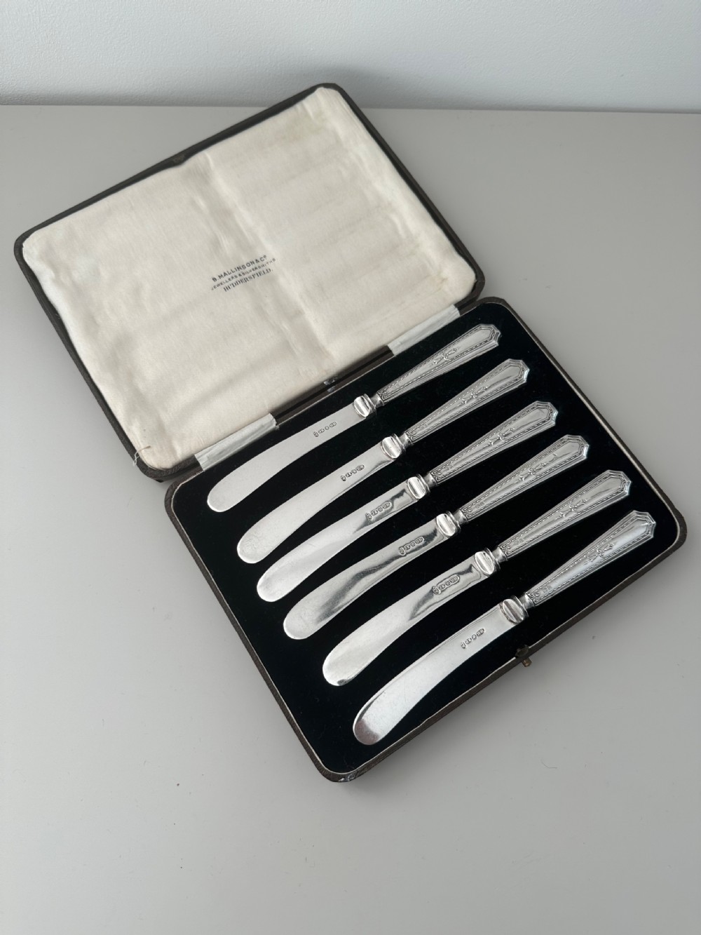 A set of six Edwardian silver butter knives with engraved handles resting inside a dark velvet-lined presentation box.
