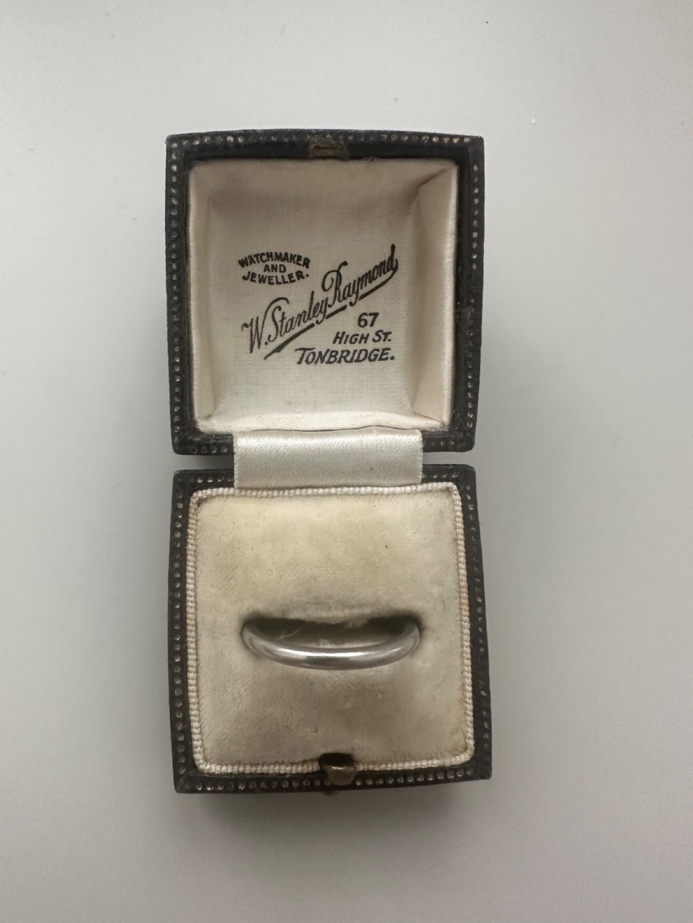 A plain platinum wedding band displayed inside an open vintage velvet-lined leather ring box. View III