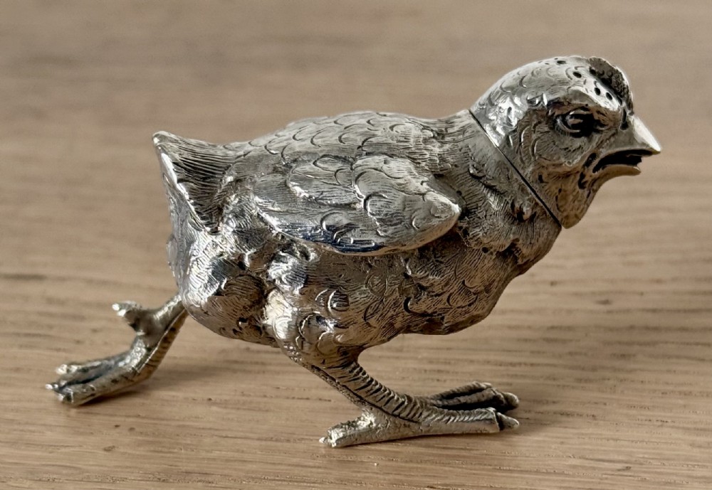 A cast silver pepper pot sculpted in the realistic shape of a chick with detailed feather engraving and a pierced head.