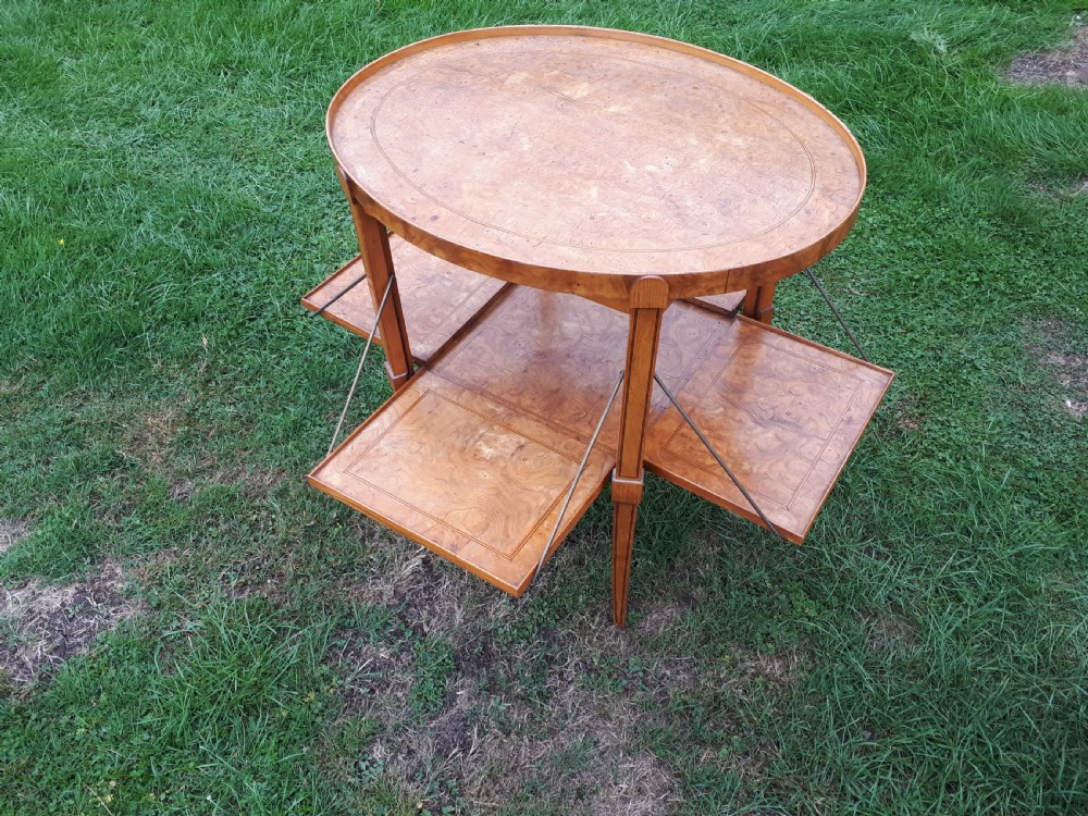 An oval golden brown burr walnut tea table featuring four extendable fold-out tray leaves supported by metal rods and standing on square tapered legs.