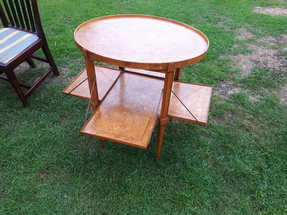 An oval golden brown burr walnut tea table featuring four extendable fold-out tray leaves supported by metal rods and standing on square tapered legs. View V