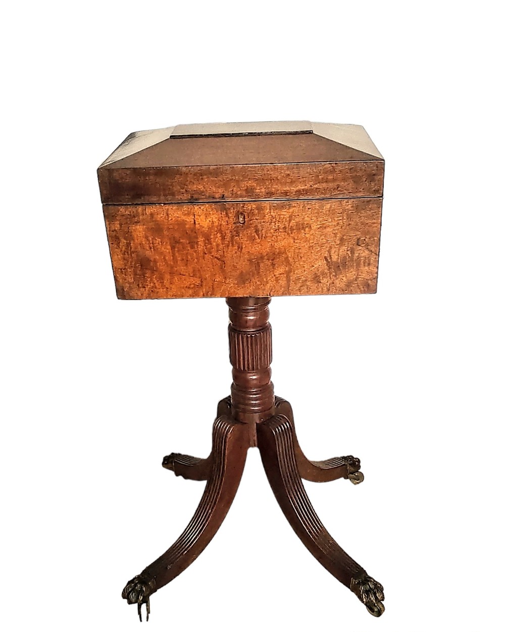 An antique mahogany teapoy featuring a hinged sarcophagus-shaped top box elevated on a reeded central pedestal with splayed legs terminating in brass paw feet.