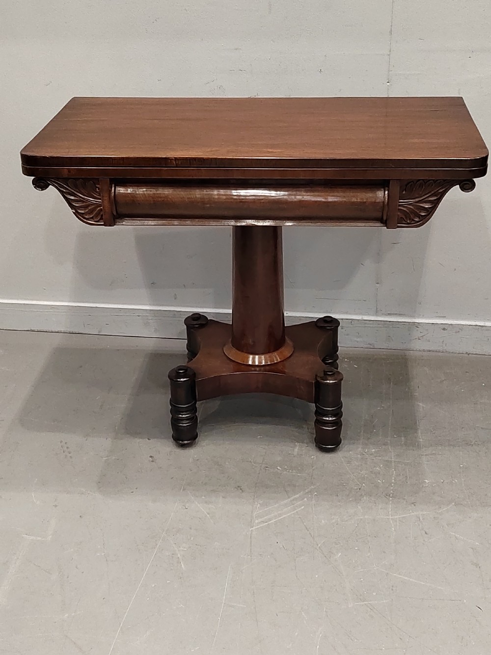 A Georgian mahogany card table featuring a hinged fold-over top, carved acanthus apron brackets, a turned pedestal column, and a shaped plinth base on turned feet.