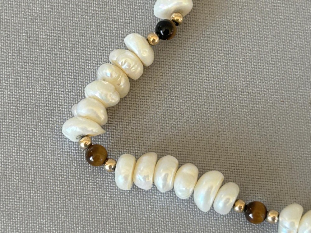 A 9ct gold strand necklace composed of irregular off-white freshwater pearls interspersed with small spherical brown tigers eye beads. View IV