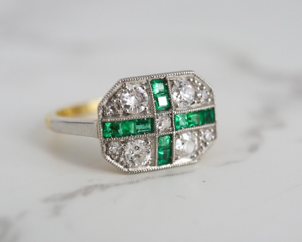 An Art Deco rectangular plaque ring featuring a cross motif of green emeralds surrounded by four round diamonds set in platinum on a gold band.