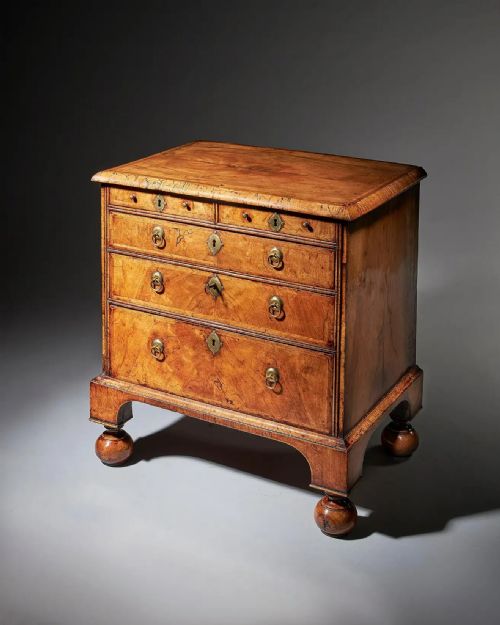fig 1. A George I figured walnut chest of drawers featuring brass drop handles, keyhole escutcheons, and a moulded base resting on turned ball and bracket feet.