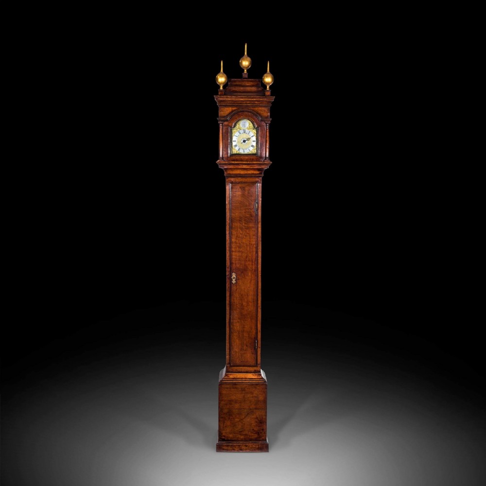 A tall Georgian longcase clock crafted from rich brown oak, featuring a brass dial, arched hood, and three brass ball finials.