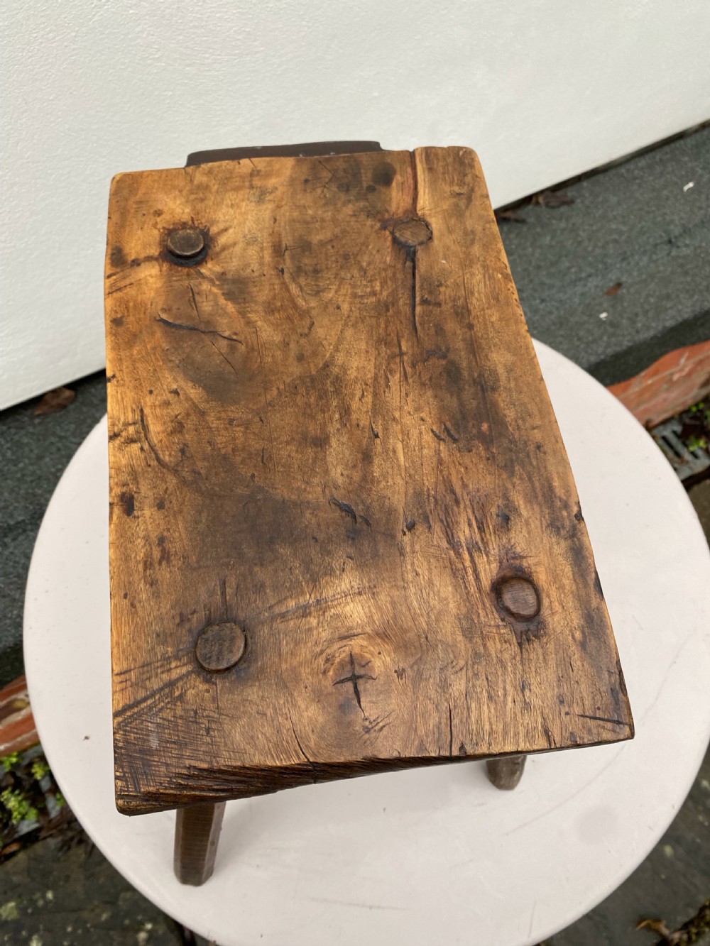 A rustic hand-carved rectangular wooden stool with four tapered legs wedged through a thick solid timber seat. View II