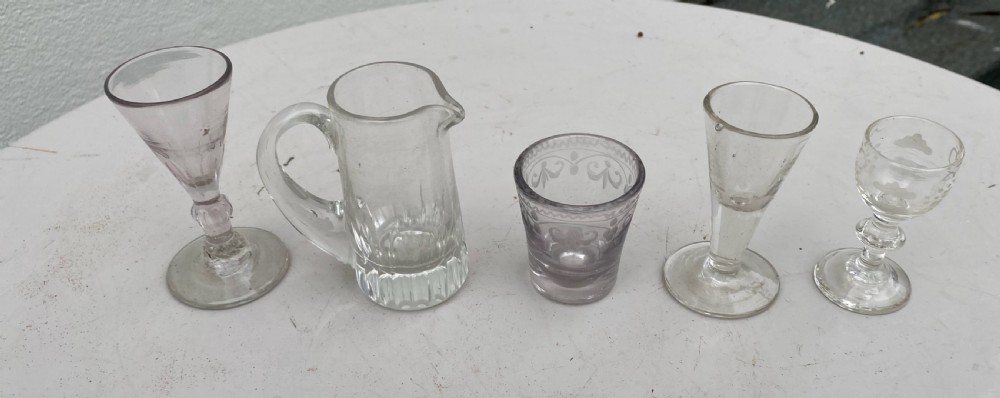 A set of five miniature glass vessels including stemmed cordial glasses, an etched tumbler, and a small jug lined up on a white surface.