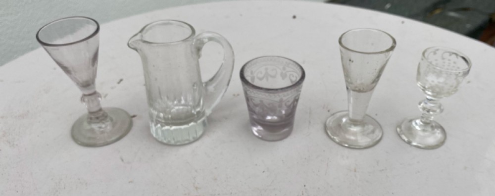 A set of five miniature glass vessels including stemmed cordial glasses, an etched tumbler, and a small jug lined up on a white surface. View II