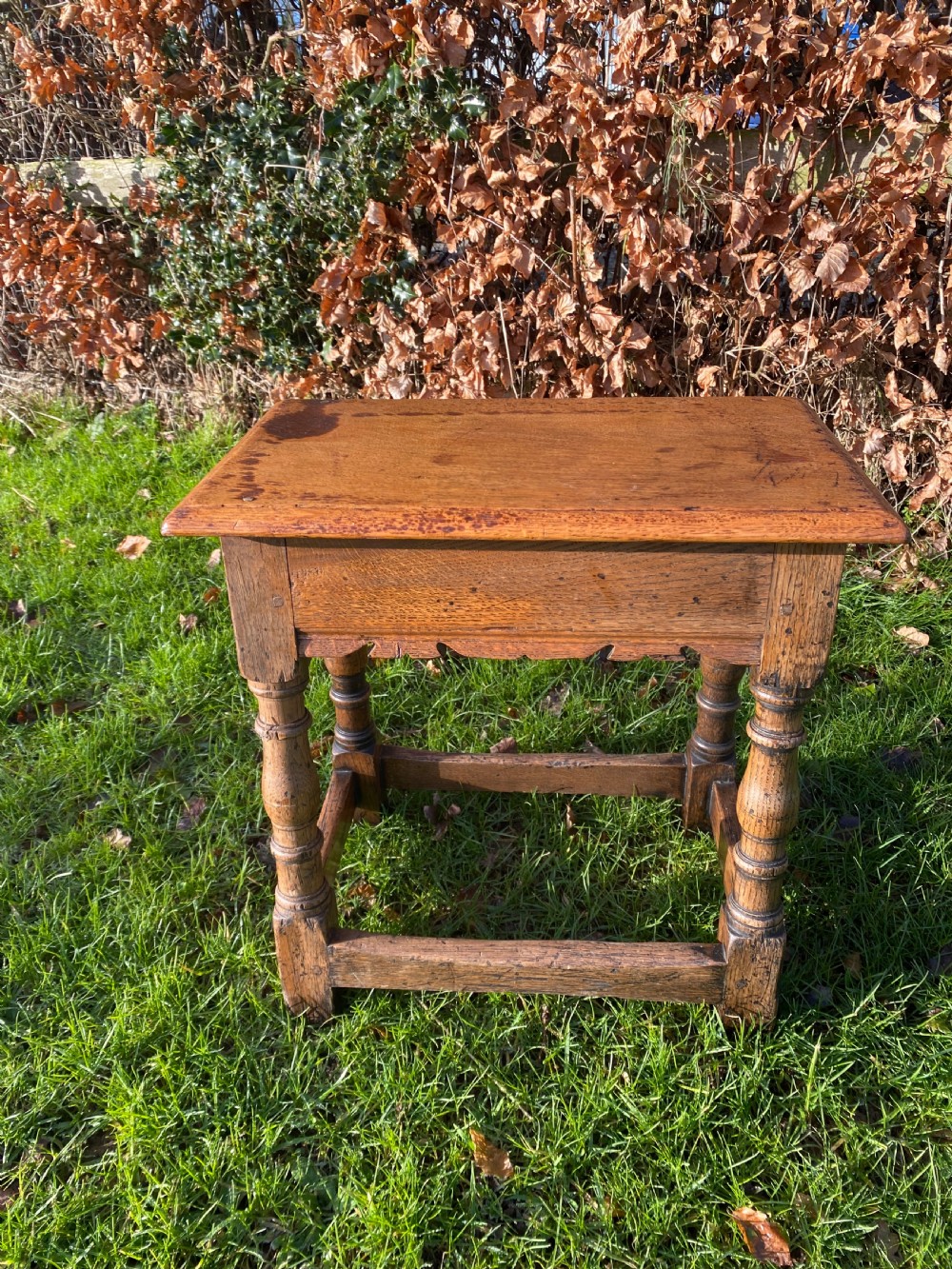 A solid oak joint stool featuring turned legs, lower perimeter stretchers, a shaped frieze apron, and a rectangular seat top.