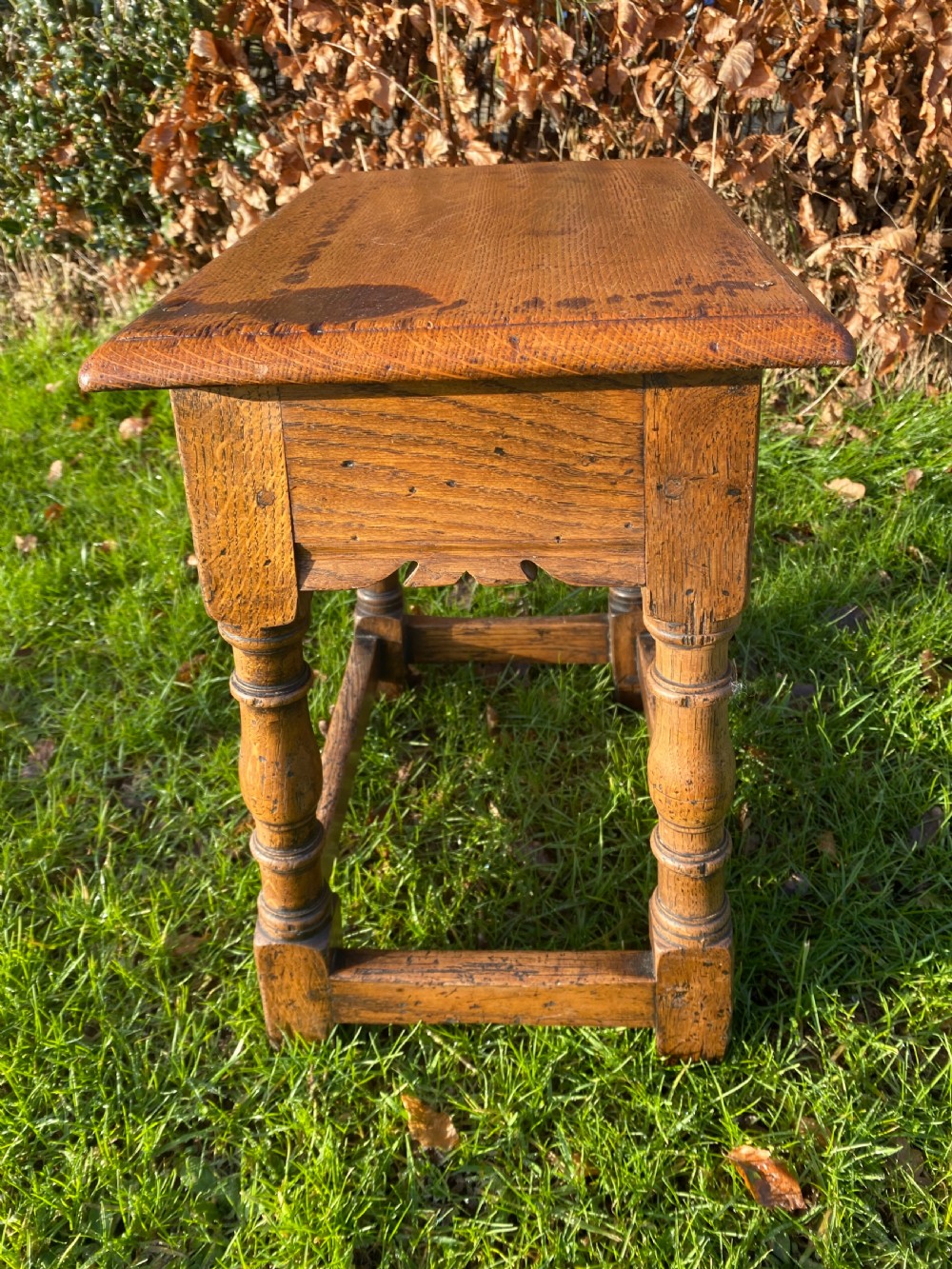 A solid oak joint stool featuring turned legs, lower perimeter stretchers, a shaped frieze apron, and a rectangular seat top. View II