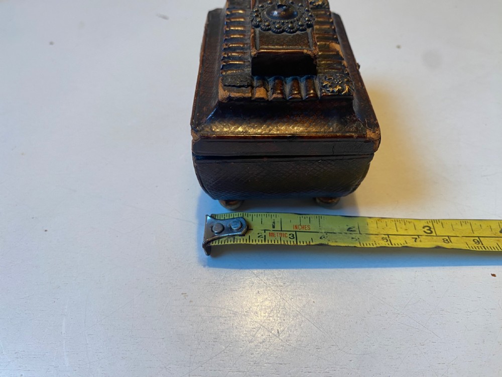 An antique dark brown tooled leather trinket box featuring a tiered lid with decorative brass mounts, brass hook clasp, and round ball feet. View XIV