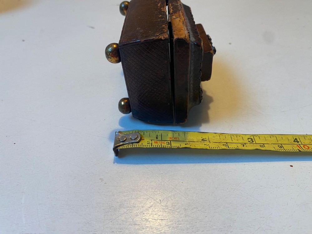 An antique dark brown tooled leather trinket box featuring a tiered lid with decorative brass mounts, brass hook clasp, and round ball feet. View XV
