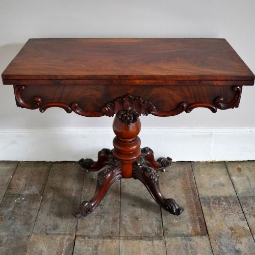 fig 2. A Victorian figured mahogany card table featuring a turnover top, carved frieze, turned pedestal, and scroll legs with castors.
