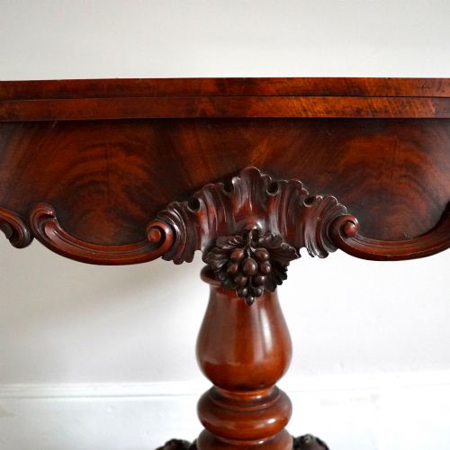 fig 5. A Victorian figured mahogany card table featuring a turnover top, carved frieze, turned pedestal, and scroll legs with castors.