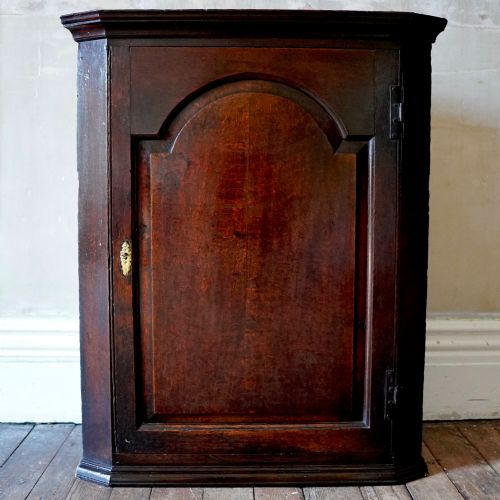 fig 1. A dark oak George III wall-hanging corner cupboard featuring an arched fielded panel door, moulded cornice, and brass escutcheon.