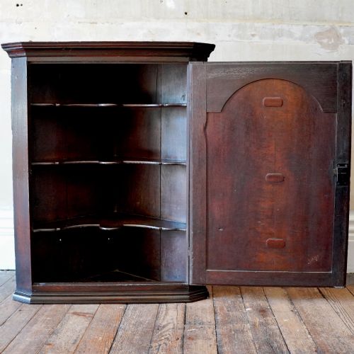 fig 2. A dark oak George III wall-hanging corner cupboard featuring an arched fielded panel door, moulded cornice, and brass escutcheon.