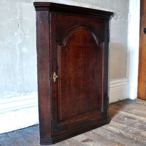 fig 3. A dark oak George III wall-hanging corner cupboard featuring an arched fielded panel door, moulded cornice, and brass escutcheon.