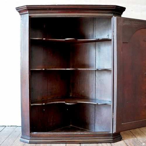fig 6. A dark oak George III wall-hanging corner cupboard featuring an arched fielded panel door, moulded cornice, and brass escutcheon.