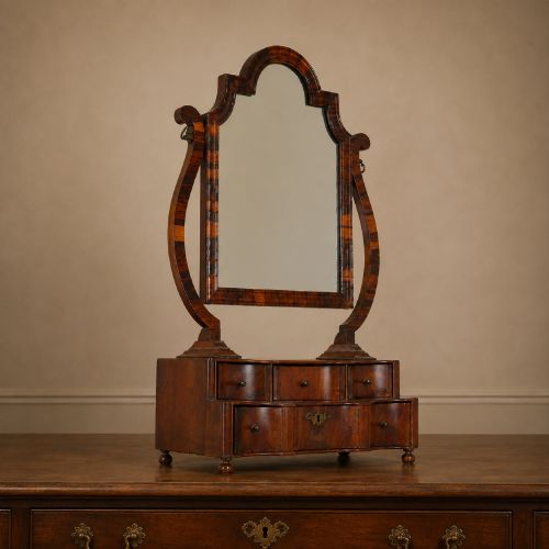 fig 1. An antique Queen Anne walnut and laburnum toilet mirror featuring an arched cross-banded frame above a serpentine base with miniature drawers.