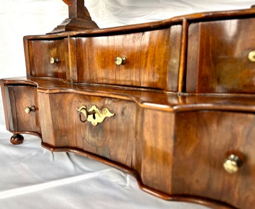 fig 3. An antique Queen Anne walnut and laburnum toilet mirror featuring an arched cross-banded frame above a serpentine base with miniature drawers.