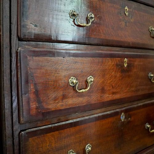 fig 6. A Georgian dark oak chest of drawers featuring two short drawers over three long drawers with mahogany crossbanding and brass swan-neck handles.