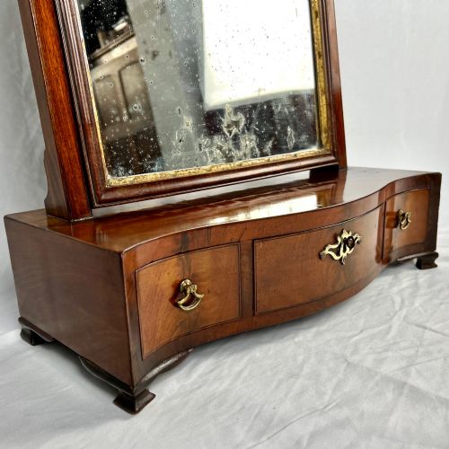 fig 3. A George III mahogany toilet dressing mirror featuring an upright rectangular swing mirror flanked by brass finials, mounted on a serpentine-front base with three small drawers.