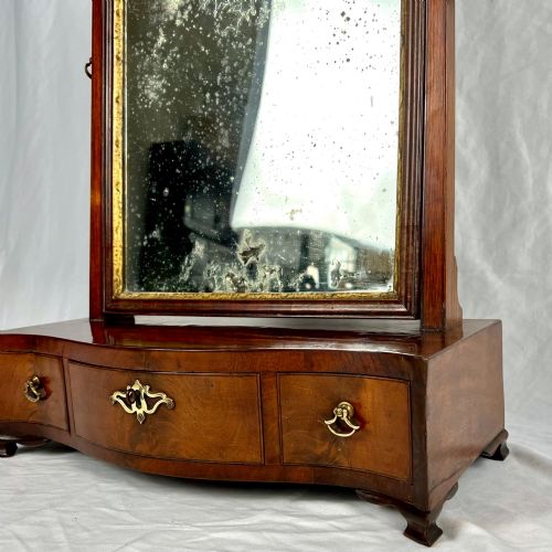 fig 4. A George III mahogany toilet dressing mirror featuring an upright rectangular swing mirror flanked by brass finials, mounted on a serpentine-front base with three small drawers.