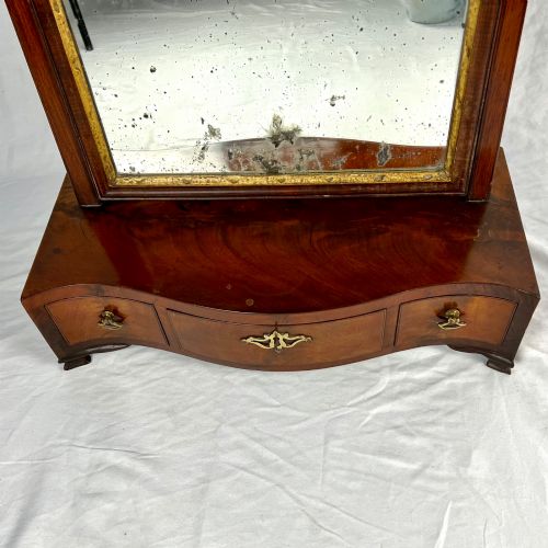 fig 5. A George III mahogany toilet dressing mirror featuring an upright rectangular swing mirror flanked by brass finials, mounted on a serpentine-front base with three small drawers.