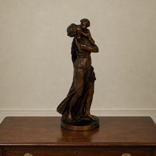 fig 6. A patinated dark bronze sculpture depicting a draped classical woman holding a small child against her face, set upon a rounded bronze base.