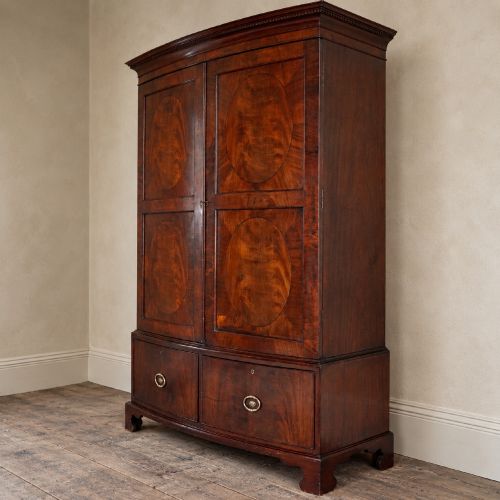 fig 3. A George III bowfront mahogany wardrobe featuring twin panelled doors with oval flame mahogany veneers above two drawers and flared bracket feet.