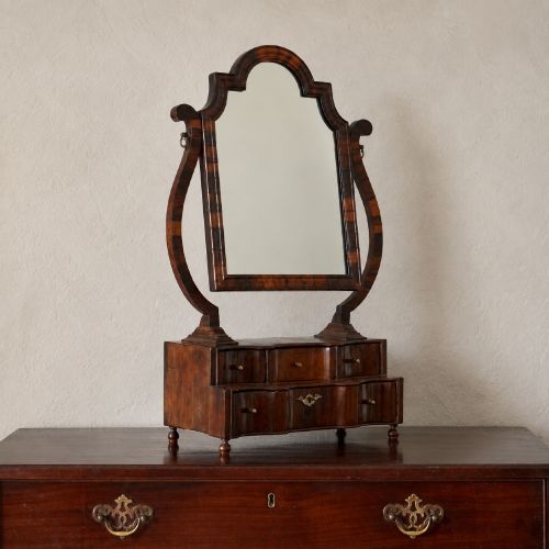 fig 1. An antique Queen Anne walnut and laburnum toilet mirror featuring an arched cross-banded frame above a serpentine base with miniature drawers.