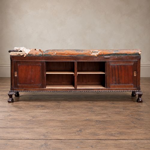 fig 2. A long antique dark mahogany bench with four panelled cupboard doors, carved ball and claw feet, and a worn leather cushion top with a raised headrest.