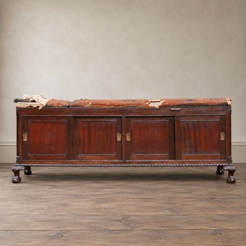 fig 3. A long antique dark mahogany bench with four panelled cupboard doors, carved ball and claw feet, and a worn leather cushion top with a raised headrest.