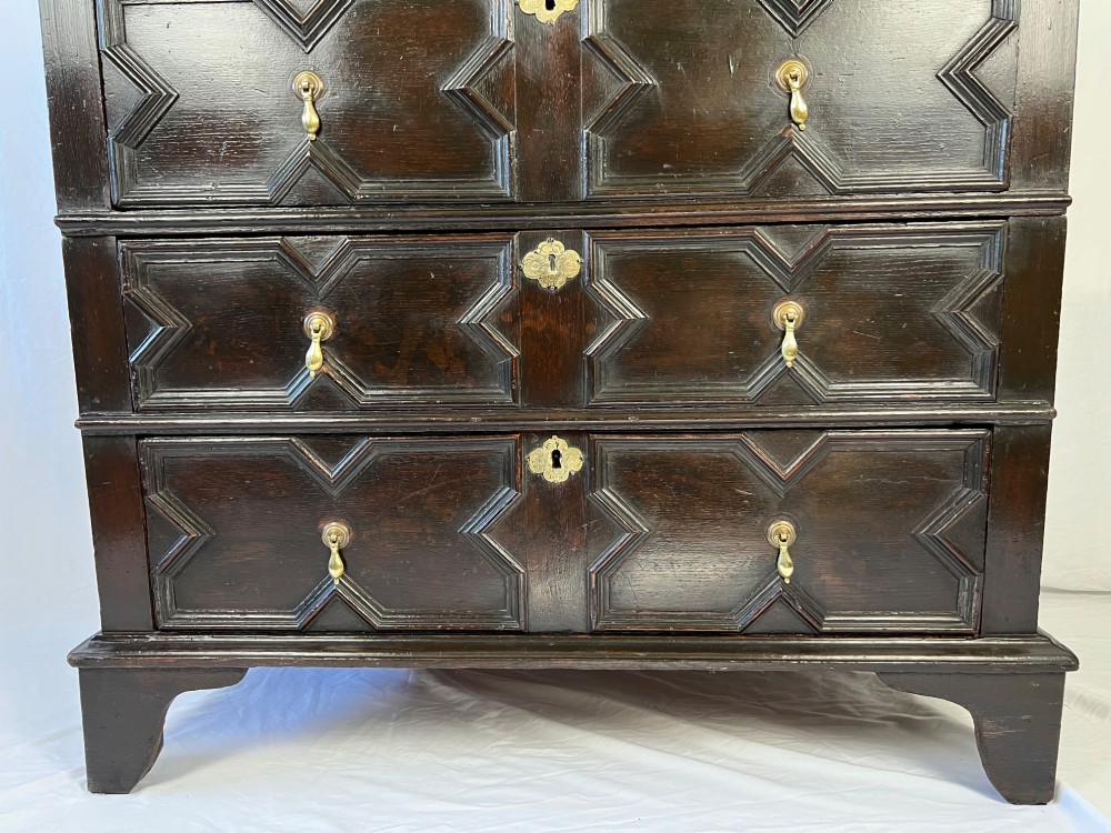 A dark oak Charles II chest of drawers featuring four geometric moulded drawer fronts with brass teardrop handles and decorative brass escutcheons. View II