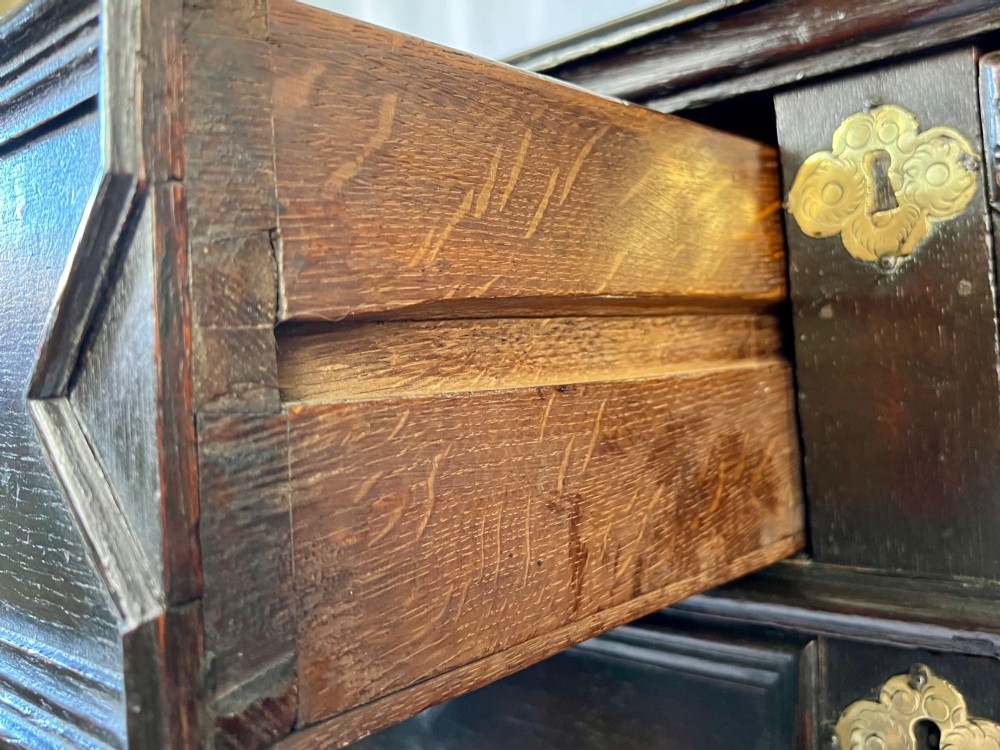 A dark oak Charles II chest of drawers featuring four geometric moulded drawer fronts with brass teardrop handles and decorative brass escutcheons. View IX