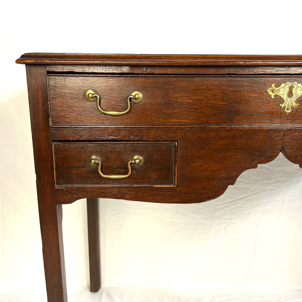 A George III dark oak lowboy table featuring three drawers with brass swan-neck handles, a shaped apron, and four square straight legs. View II