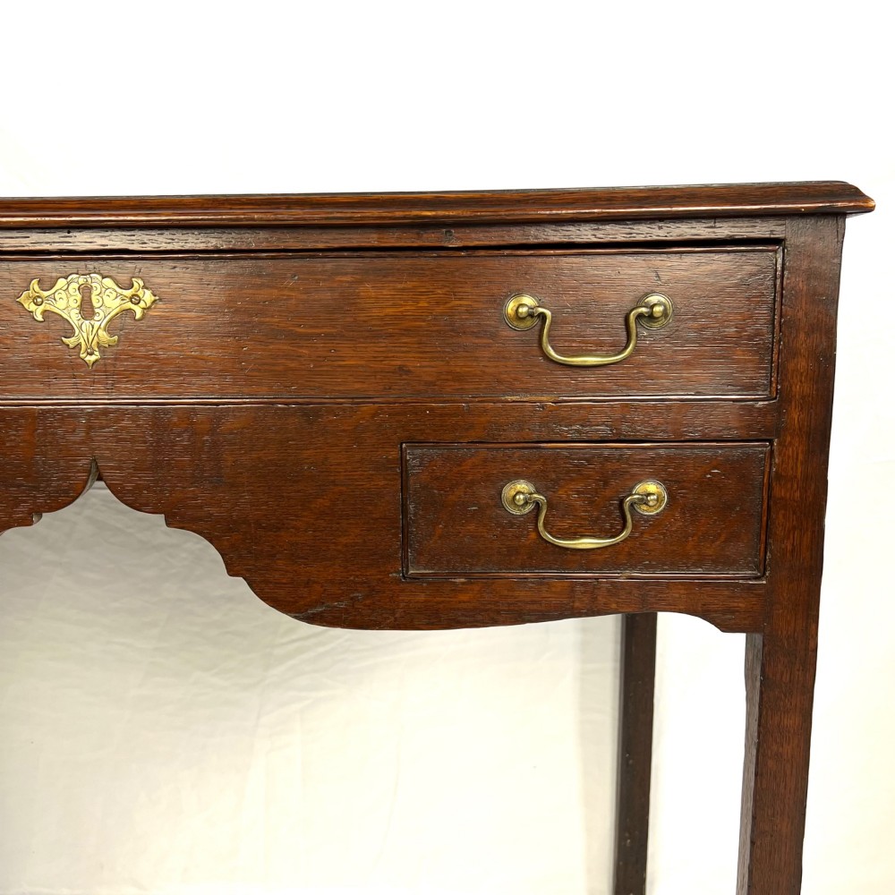 A George III dark oak lowboy table featuring three drawers with brass swan-neck handles, a shaped apron, and four square straight legs. View IV