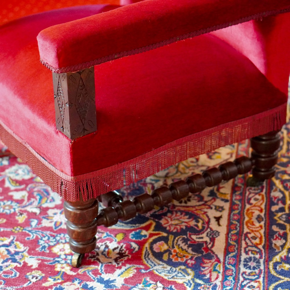 A pair of oversized Victorian armchairs upholstered in crimson red velvet with fringe trim, featuring carved oak arm supports and bobbin-turned legs. View X
