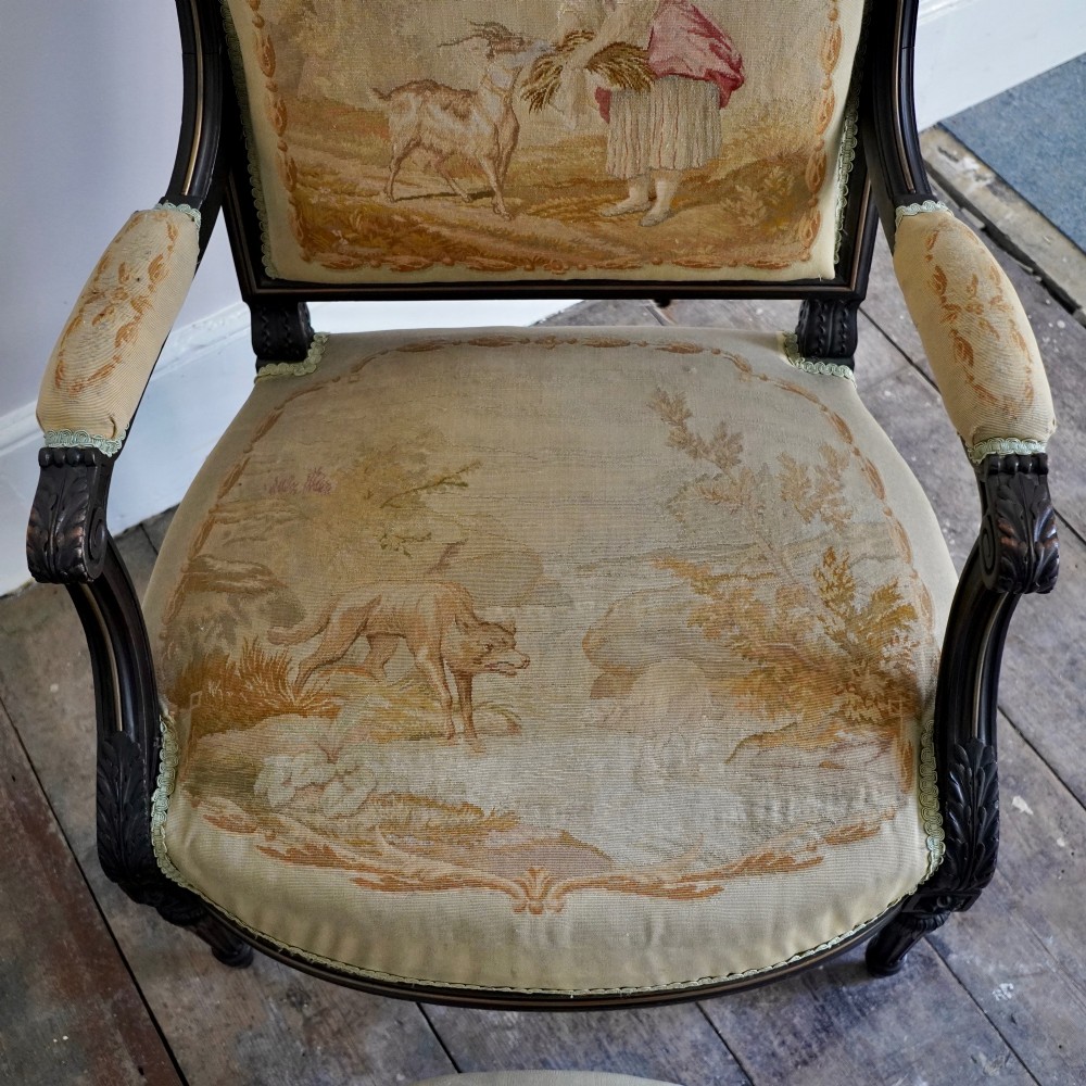 A pair of ebonised beech Louis XVI Revival salon armchairs featuring carved frames, fluted legs, padded armrests, and tapestry upholstery, accompanied by matching footstools. View V