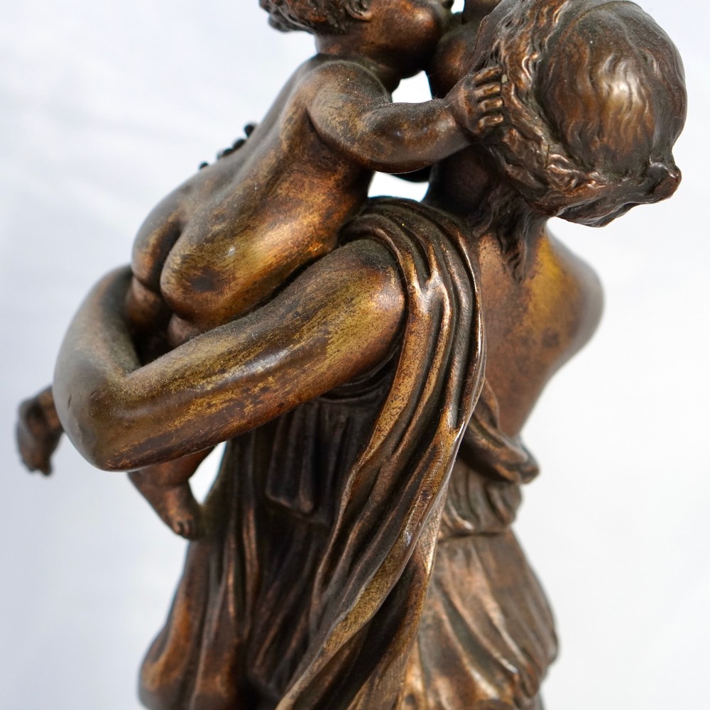 A patinated dark bronze sculpture depicting a draped classical woman holding a small child against her face, set upon a rounded bronze base. View II