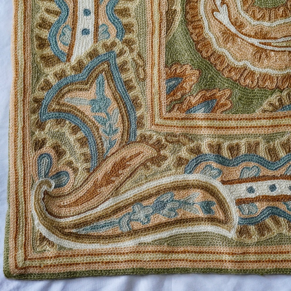 A rectangular crewelwork embroidered textile panel featuring large paisley motifs in terracotta, blue, and cream on a sage green background with a decorative border. View II