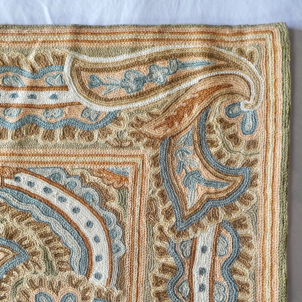 A rectangular crewelwork embroidered textile panel featuring large paisley motifs in terracotta, blue, and cream on a sage green background with a decorative border. View VII