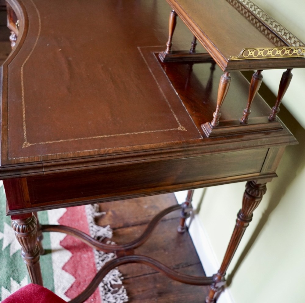 A Victorian rosewood writing desk featuring a brass gallery shelf, three drawers with brass handles, turned fluted legs, and curved stretchers. View X