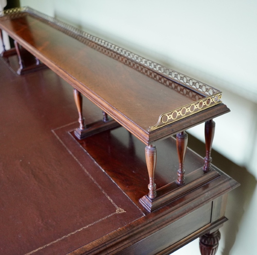 A Victorian rosewood writing desk featuring a brass gallery shelf, three drawers with brass handles, turned fluted legs, and curved stretchers. View XI